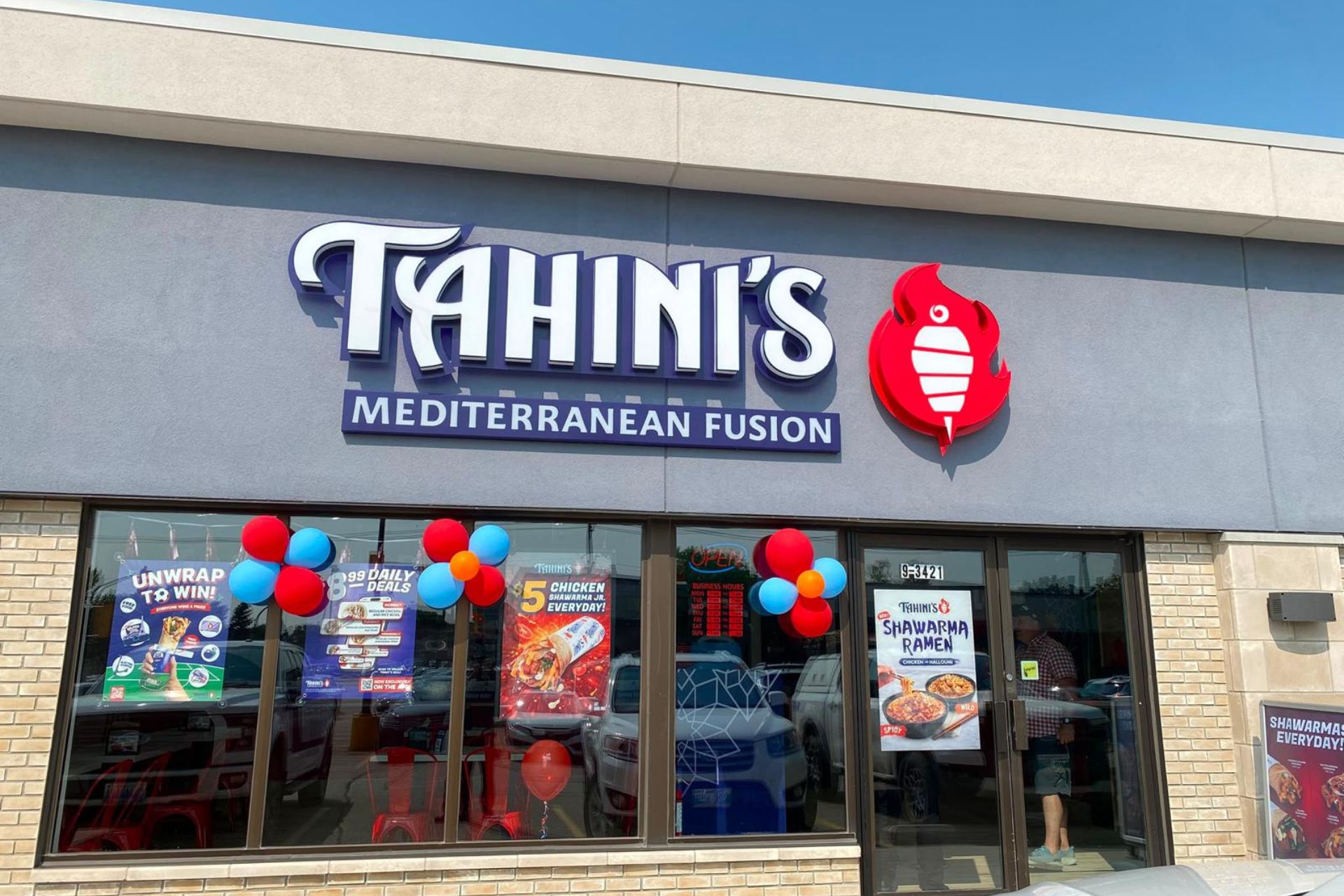 Exterior view of a Tahini's Mediterranean Fusion restaurant location in Winnipeg decorated with red and blue balloon clusters along the large front windows and glass entrance doors.